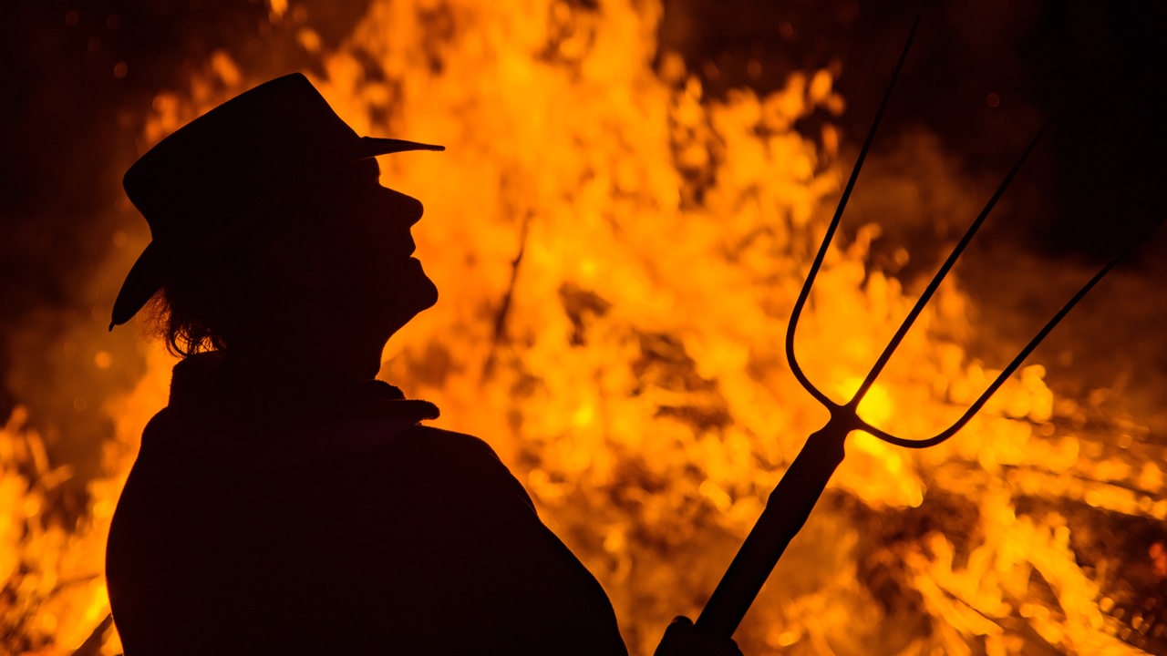 Ein Mann mit Mistgabel steht vor einem brennenden Lagerfeuer.