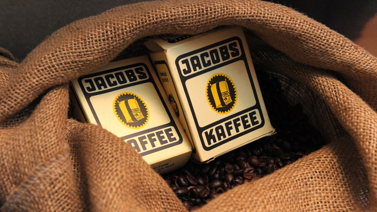 Zwei Packungen Jacobs-Kaffee liegen in einem Jutesack voller Kaffeebohnen