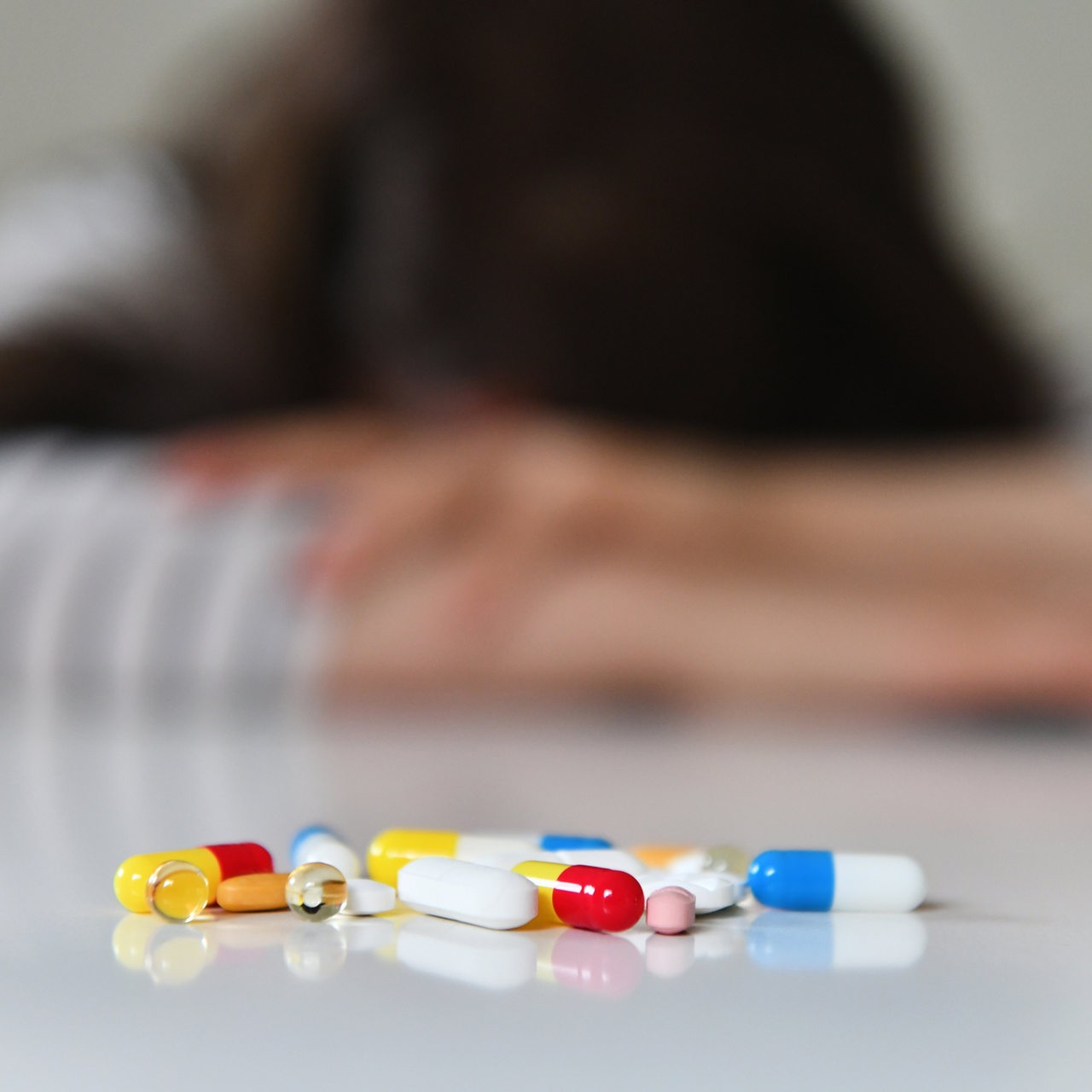 Symbolfoto zum Thema Depressionen: Tabletten liegen vor einer unscharfen Person, die ihren Kopf auf den Tisch legt.