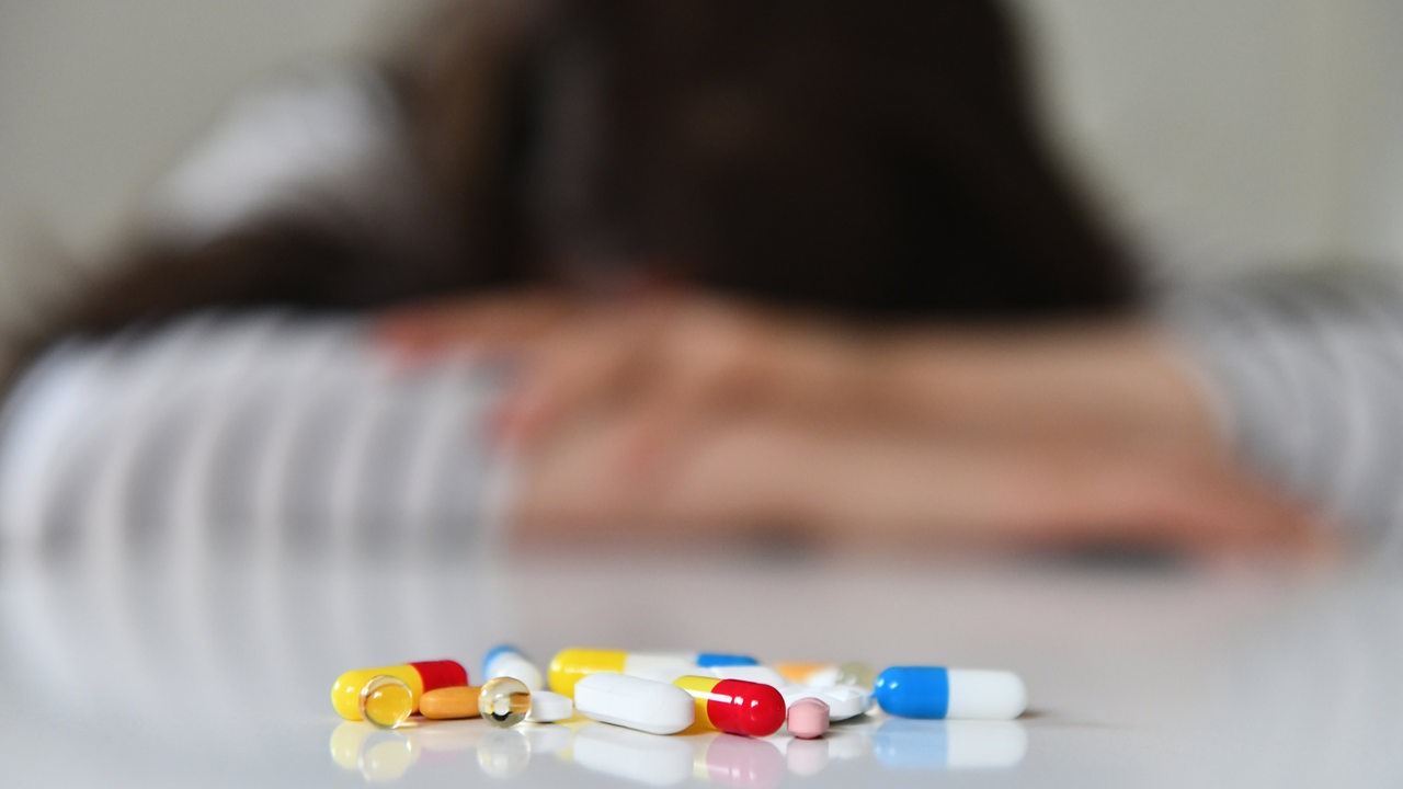 Symbolfoto zum Thema Depressionen: Tabletten liegen vor einer unscharfen Person, die ihren Kopf auf den Tisch legt.