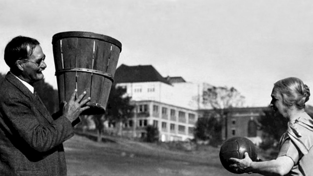Das historische Bild zeigt den kanadischen Sportlehrer Dr. James Naismith im Jahr 1891 in den USA. Er hält einen Korb in den eine ältere Frau einen Ball werfen soll