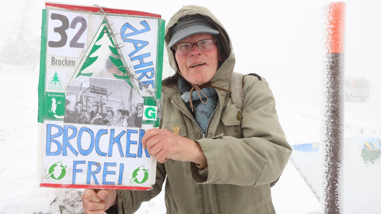 Wanderer Benno Schmidt, besser bekannt als Brocken Benno, steht mit einem Schild zur Erinnerung an die Brockenöffnung am 3.12.1989 auf dem Brocken. 