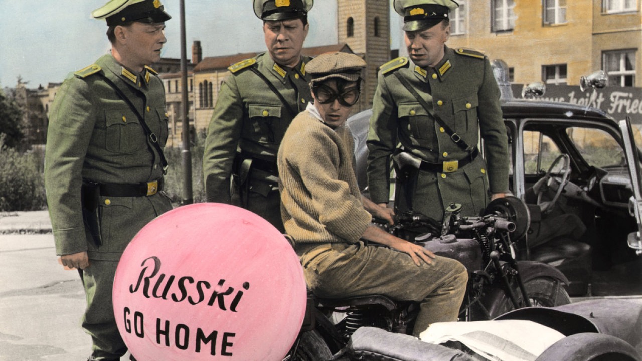 Filmszene aus Billy Wilders "Eins, Zwei,Drei", an einem Motorrad bläst sich ein Luftballon mit der Aufschrift: Russki Go Home auf, das am Grenzübergang in Berlin kontrolliert wird