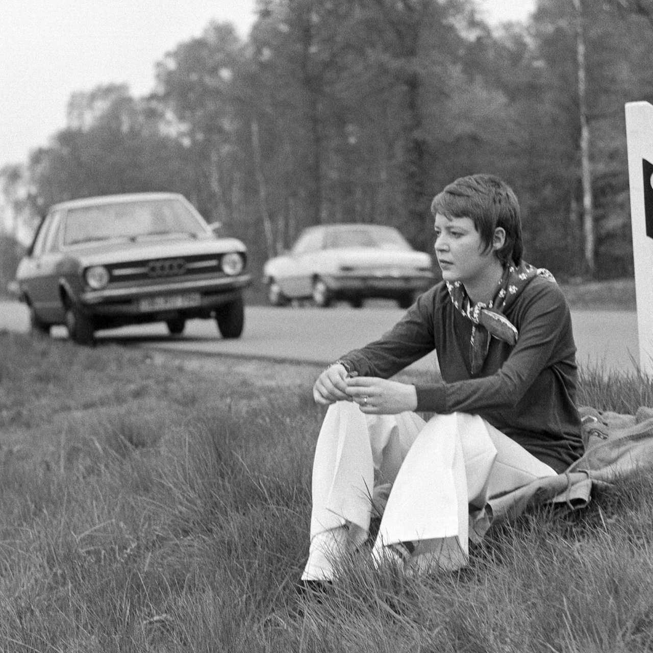 Siebziger Jahre: eine junge Frau sitzt am Strassenrand einer Landstrasse, Fahrpause, Ruhepause, im Hintergrund das geparkte Auto