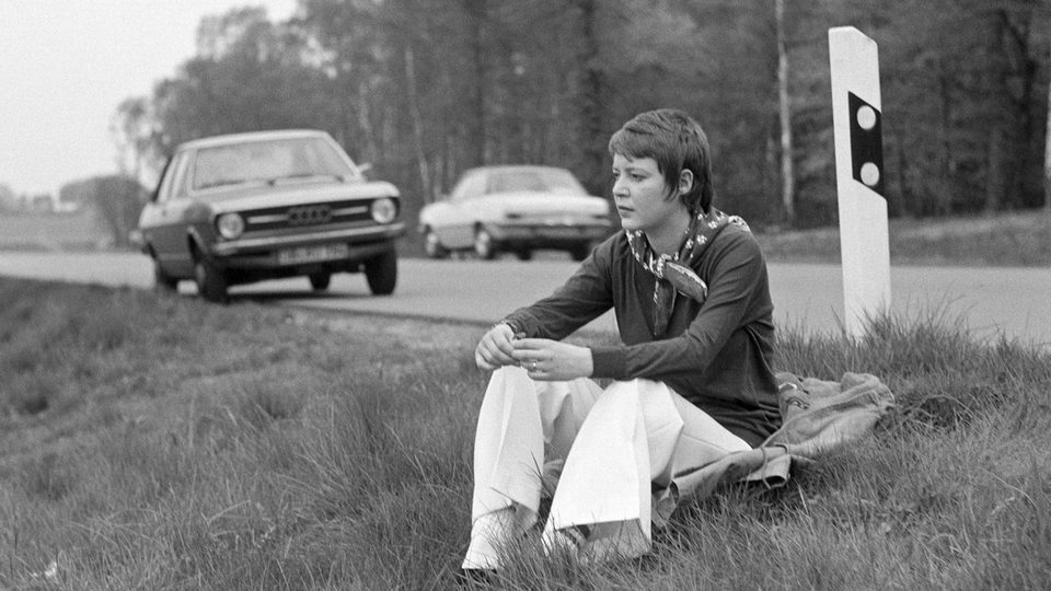 Siebziger Jahre: eine junge Frau sitzt am Strassenrand einer Landstrasse, Fahrpause, Ruhepause, im Hintergrund das geparkte Auto