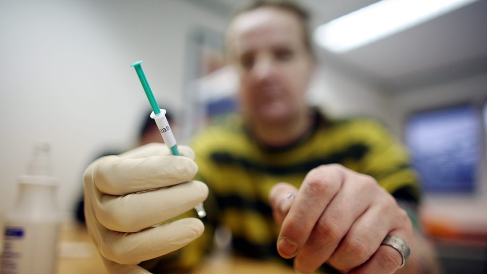 Ein Mann (unscharf) hält in einer Heroinabgabestelle mit Schutzhandschuhen eine Spritze mit Heroin in die Kamera.