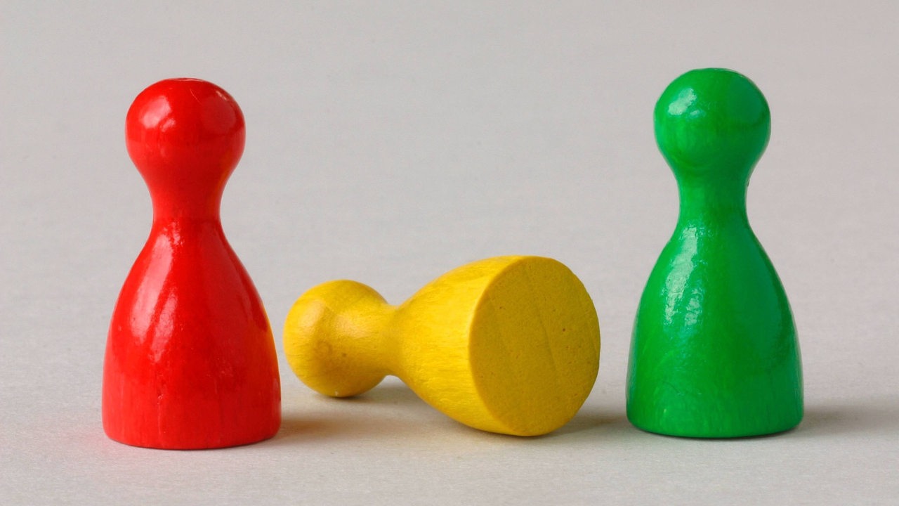 Mensch-ärgere-dich-nicht-Spielfiguren in rot, gelb und grün. Die gelbe Figur ist umgekippt.