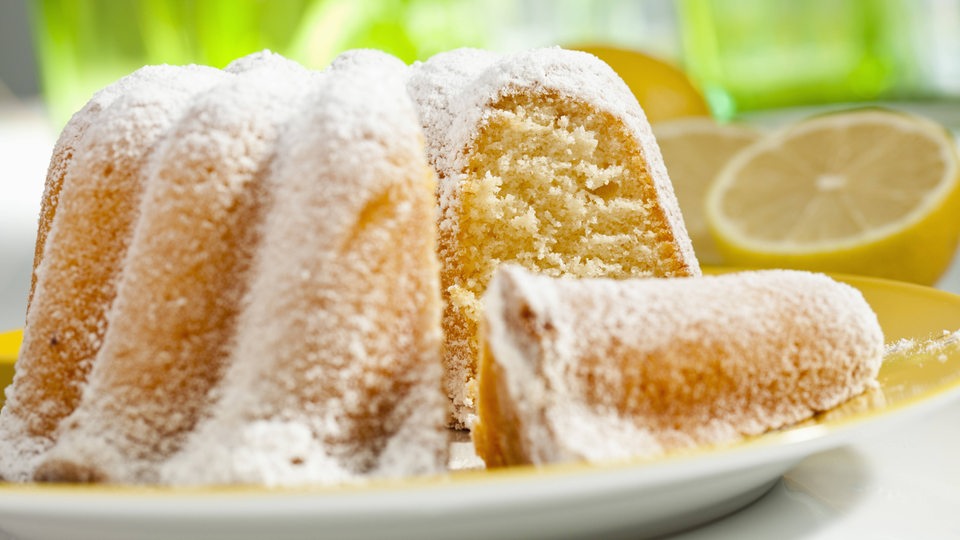 Ein saftiger Buttermilch-Zitronen-Topkuchen auf einem gelben Teller. Im Hintergrund lieg eine aufgeschnittene Zitrone. 