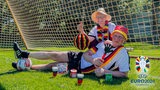 Bremen Eins Moderator:innen Katharina Guleikoff und Philipp Kolanghis in voller Euro-Fanmontur liegen vor einem Fußballtor.