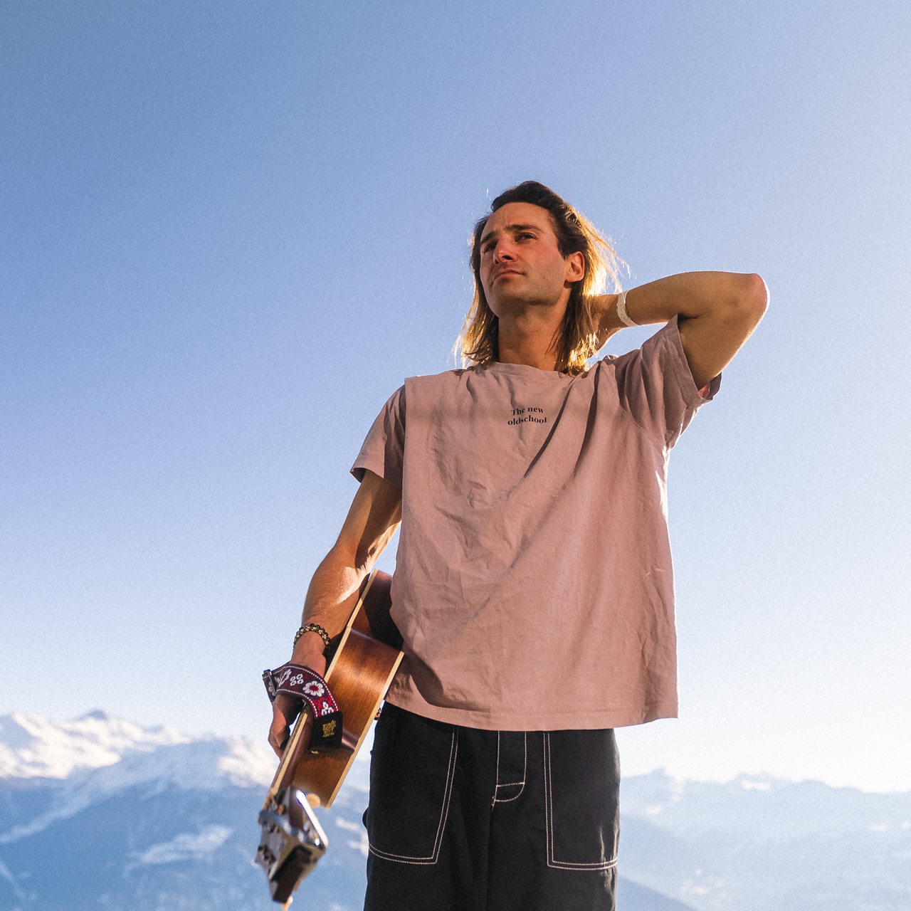 Der Musiker Pat Burgener steht mit Gitarre und T-Shirt in den Schweizer Alpen
