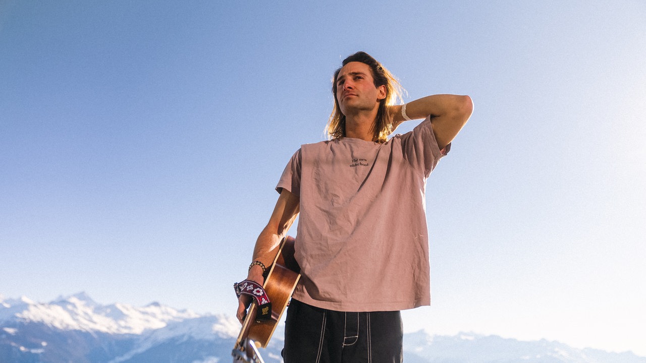 Der Musiker Pat Burgener steht mit Gitarre und T-Shirt in den Schweizer Alpen