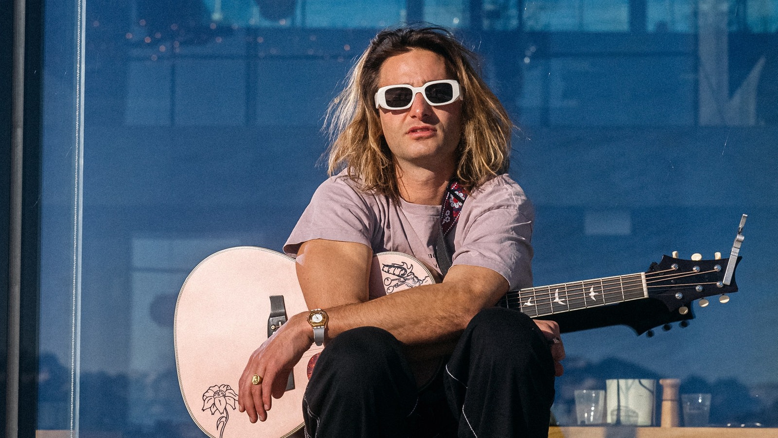Pat Burgener mit Sonnenbrille und Gitarre sitzt auf einer Holzbank vor einem Panoramafenster