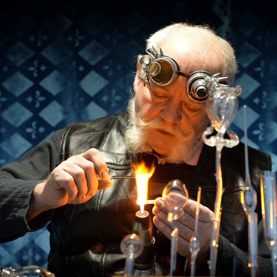 Ein Glasbläser in seiner Werkstatt hantiert mit Materialien über einer offenen Flamme.