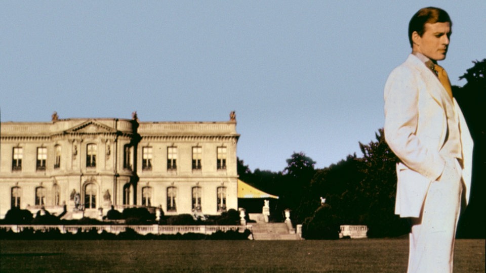 Filmszene aus "Der große Gatsby" von 1974. Robert Redford in weißem Anzug steht vor einer Villa.