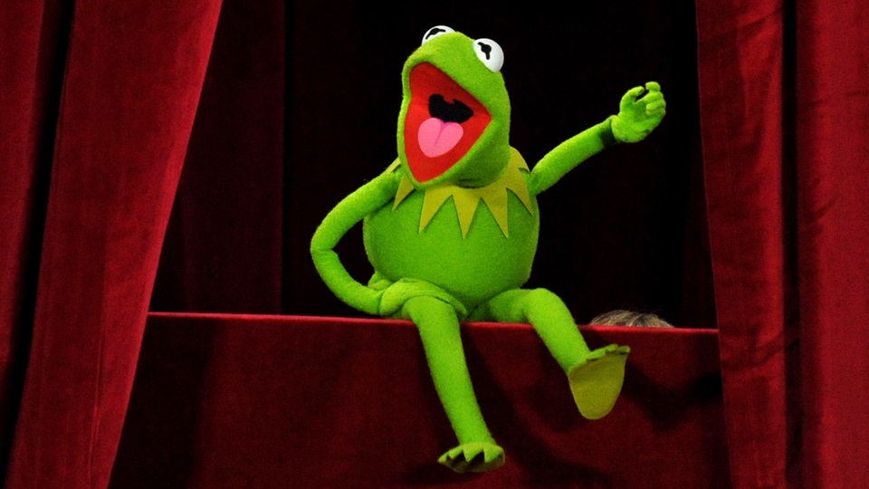 Die Puppe "Kermit der Frosch" sitzt auf der Bühne mit roten Vorhängen an der Seite.