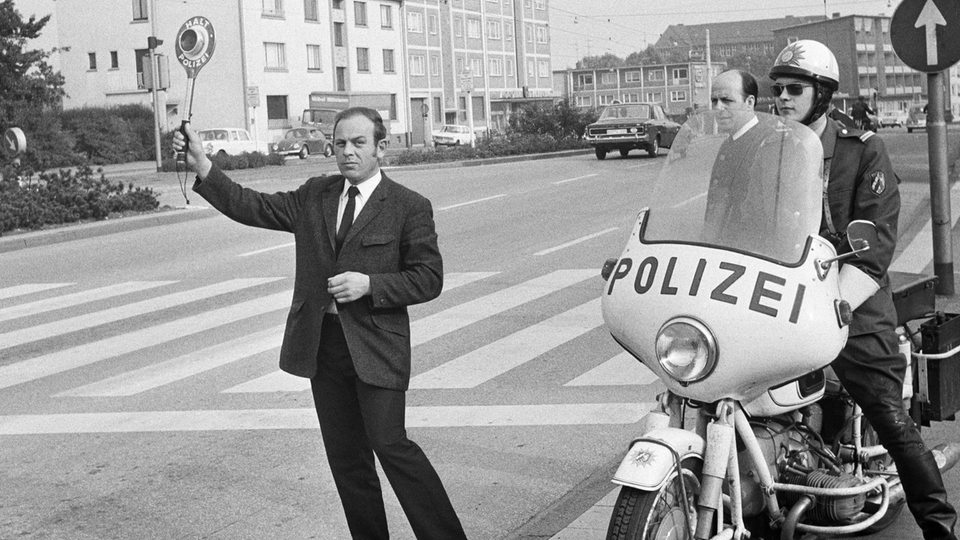 Siebziger Jahre, Schwarzweissfoto: An einem Zebrastreifen hebt ein Beamter eine Polizeikelle, daneben ein Polizist auf einem Motorrad.