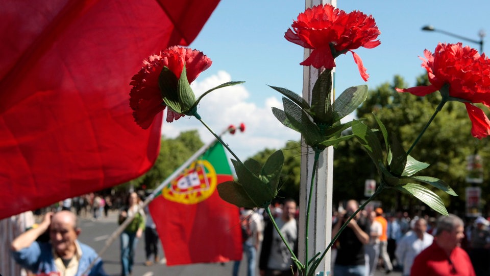 Rote Nelken im Vordergrund, im  Hintergrund ein Demonstration mit portugisischen Fahnen, die anlässlich des Jahrestages der Nelkenrevolution stattgefunden hat.