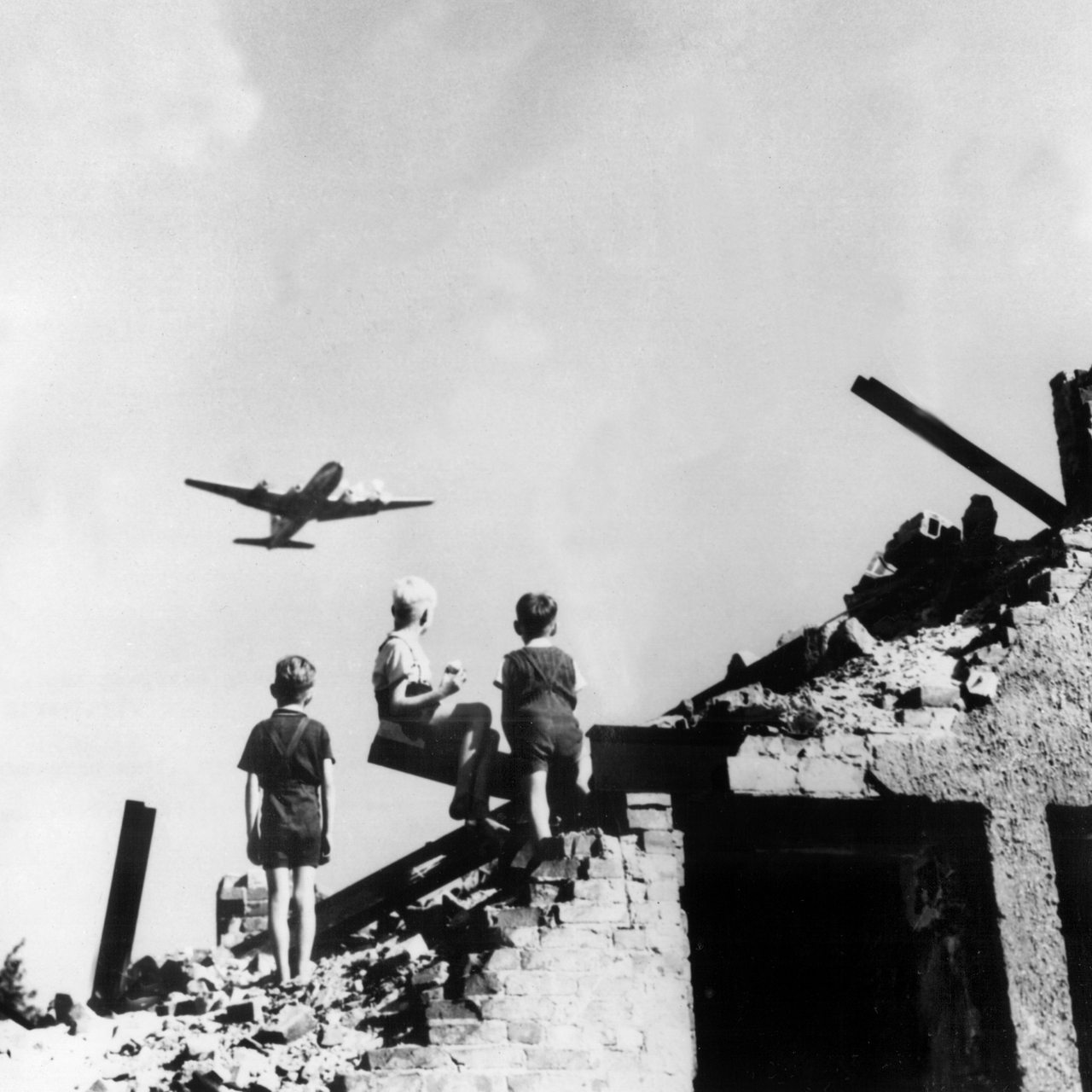 Drei West-Berliner Jungen, die auf einem Trümmerberg spielen, schauen zu einem US-amerikanisches Transportflugzeug vom Typ C-54 empor, das Versorgungsgüter nach West-Berlin bringt 