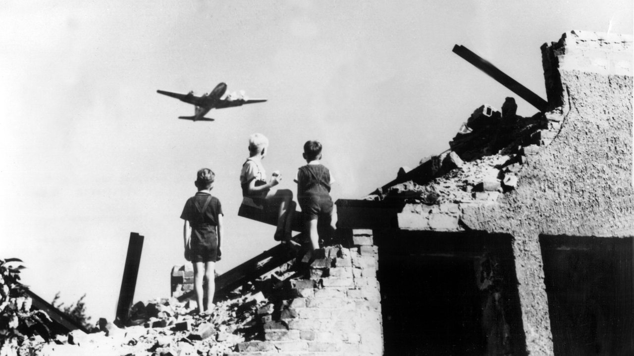 Drei West-Berliner Jungen, die auf einem Trümmerberg spielen, schauen zu einem US-amerikanisches Transportflugzeug vom Typ C-54 empor, das Versorgungsgüter nach West-Berlin bringt 