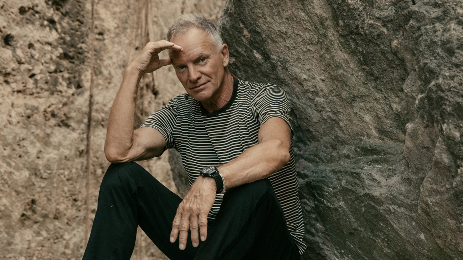 Sänger Sting trägt ein gestreiftes Shirt und eine schwarze Jeans mit schwarzen Boots und sitzt auf einem Stein