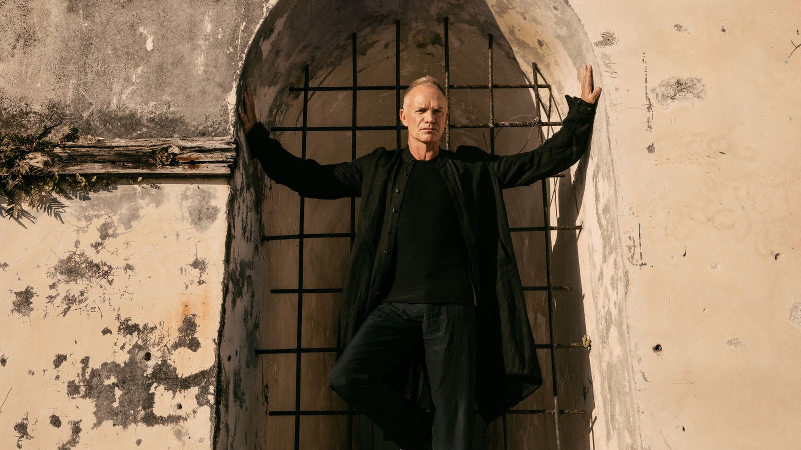 Sänger Sting trägt ein schwarzes Shirt, eine schwarze Jeans und einen schwarzen Mantel und steht in einer Tür. Seine Hände drücken gegen die Wände rechts und links.