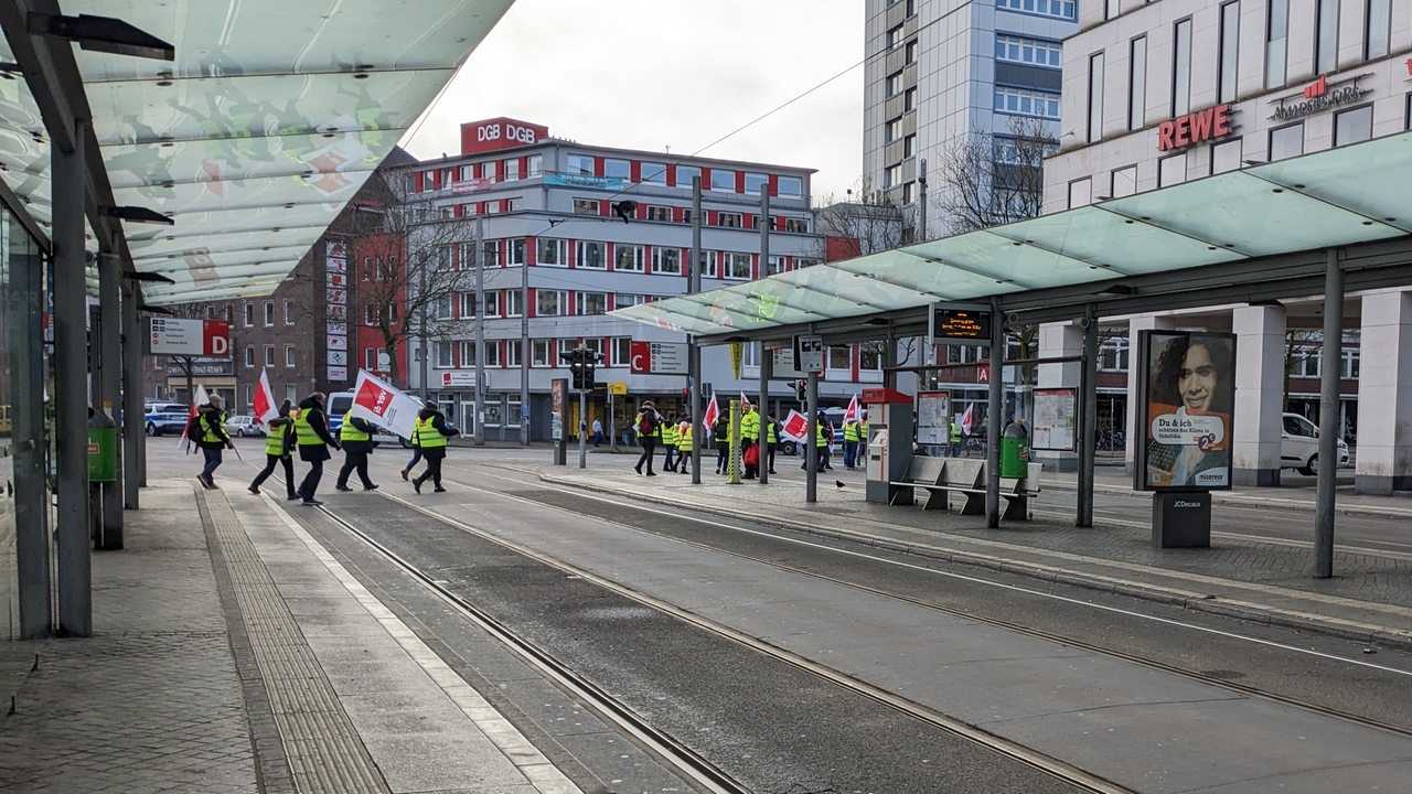 Menschen in Warnwesten und mit Verdi-Fahnen laufen auf dem Bremer Bahnhofsplatz über eine Straße.