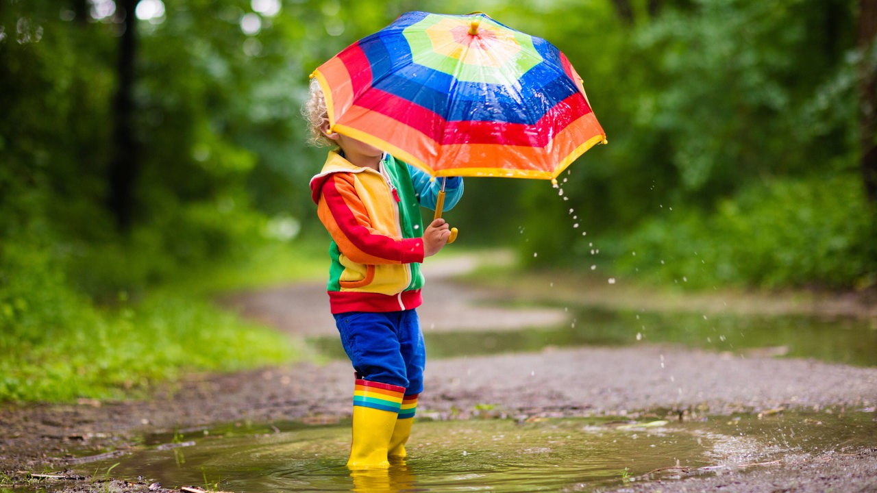 Kind in bunter Kleidung und buntem Regenschirm steht in einer Pfütze